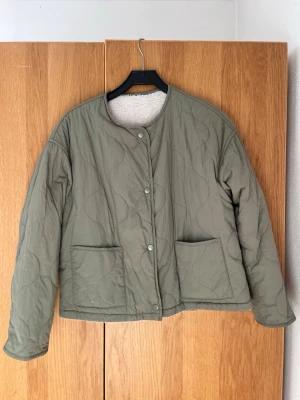 Olivgrön pufferjacka med teddyfoder - Säljer en olivgrön pufferjacka med vågigt quiltat mönster och två stora framfickor. Jackan har tryckknappar och ett mysigt vitt teddyfoder på insidan. Rak passform och rundad halsringning. Man kan ha den på båda hållen så det finns inte nått ut och in på den😊