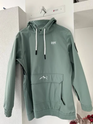 Grön hoodie från Dope Snow - Ljusgrön hoodie från Dope Snow med stor huva, vita dragsnören och en stor magficka med bergstryck. Tillverkad i slitstarkt syntetmaterial, har ribbade muddar och dragkedjor i sidorna för extra funktion. Perfekt för sport och utomhusaktiviteter.