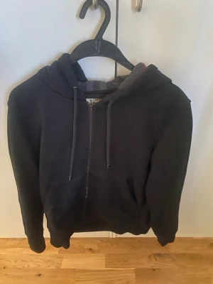 Svart burberry hoodie - Svart hoodie med dragkedja och fickor framtill. Huvan har ett rutigt innerfoder i grått och rött, samt justerbara snören. Perfekt för en avslappnad och chill stil. Tillverkad i mjukt material för extra komfort.