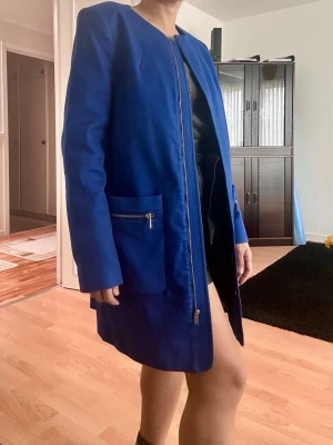 Blue coat from Mango - Snygg kornblå rock i rak modell med rund halsringning och långa ärmar. Rocken har två stora fickor framtill med silverfärgade dragkedjor och stängs med en dold dragkedja. Perfekt för dig som vill sticka ut med färg och stil.
