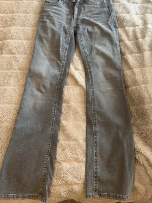 Grå bootcut jeans från Gina - Säljer ett par grå bootcut jeans med snygga fickdetaljer bak, dekorerade med mönstrat band och silverfärgade knappar. Klassisk femficksmodell och normal midja. Perfekta för dig som gillar en avslappnad men ändå trendig look.