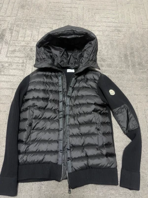 Svart pufferjacka från Moncler - Snygg svart pufferjacka från Moncler med stickade ärmar och huva. storlek M men passar även som S Jackan har två dragkedjefickor fram och Moncler-logga på ena ärmen. Perfekt för kalla dagar med sin vadderade framsida och ribbade muddar.