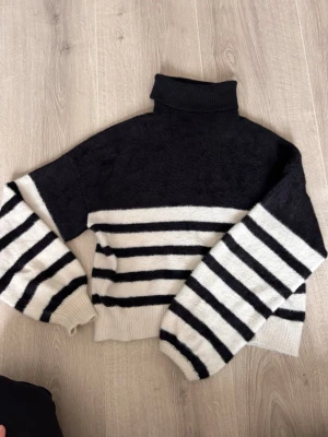 Randig svartvit polotröja - Snygg stickad polotröja med svart överdel och vita ärmar och nederdel med svarta horisontella ränder. Tröjan har en hög krage och vida ärmar, perfekt för en trendig och avslappnad look.