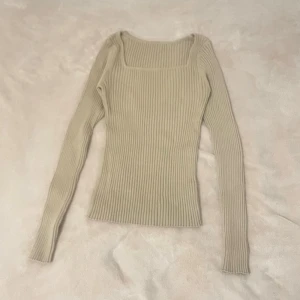 Beige ribbstickad topp med fyrkantig ringning - Snygg beige ribbstickad topp med lång ärm och trendig fyrkantig ringning. Figurnära passform och stretchigt material som ger en clean och modern look. Perfekt att styla med jeans eller kjol för en enkel men chic outfit.