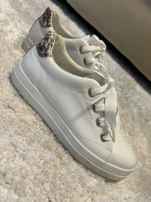 Vita sneakers från Gant med ormmönster - Snygga vita sneakers från Gant med coola ormmönstrade detaljer i beige och brunt på hälen. Skorna har vita snören, guldfärgade öljetter och en klassisk låg profil. Perfekta för dig som vill ha en clean look med en twist.