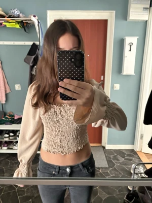 Beige smockad topp från Boohoo - Trendig beige topp från Boohoo med smockad kropp och långa puffärmar. Toppen har resår vid axlar och ärmslut för en snygg passform och volym. Perfekt croppad längd och mjukt, lätt material som känns skönt mot huden. Passar säkert M