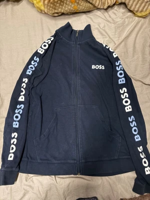 Marinblå zip hoodie från BOSS - Snygg marinblå hoodie från BOSS med dragkedja framtill och vita BOSS-loggor längs båda ärmarna. Tröjan har hög krage, två fickor framtill och ribbade muddar. Tillverkad i mjukt bomullsmaterial för en bekväm och avslappnad stil.