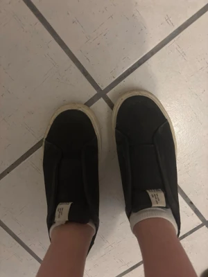 Svarta skor - Säljer ett par svarta slip-on sneakers från lager med vit sula och minimalistisk design. Skorna har en bred elastisk panel över foten och är tillverkade i canvas. Perfekta för dig som gillar stilrena och bekväma sneakers.