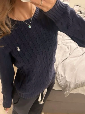 Mörkblå kabelstickad tröja Polo Ralph Lauren - Mörkblå kabelstickad tröja från Polo Ralph Lauren med rund halsringning och vit broderad logga på bröstet. ’’’ först bild är inte min men det är samma tröja jag säljer, har inte så bra ljus men så ser den ut på någon. Det är bara att skiva om man vill ha fler bilder. 
