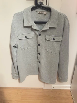 Ljusgrå overshirt från Only & Sons - Stilren ljusgrå overshirt från Only & Sons med klassisk krage och svarta knappar framtill. Två stora bröstfickor med lock och knapp. Tillverkad i mjukt, strukturerat material som ger en avslappnad look. Perfekt att bära öppen eller stängd.