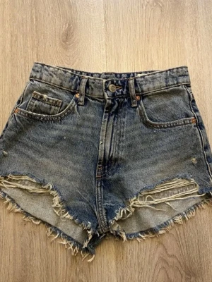 Jeansshorts - Säljer ett par ljus-blå jeansshorts med lite fransiga kanter och slitna detaljer framtill. Klassisk femficksmodell med knapp och dragkedja. Perfekta för sommaren! Skriv om du har frågor💗