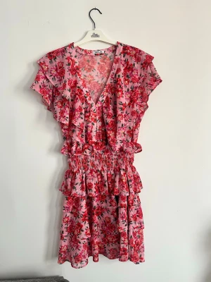 Blommig klänning  - Supersöt klänning från Replay i rosa med blommigt mönster i rött och grönt. Klänningen har volangdetaljer, v-ringning och korta ärmar. Perfekt för sommarens alla tillfällen. 