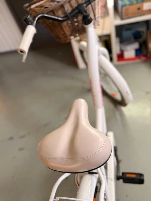 Vit damcykel med korg Goetze - Säljer en vit damcykel från Goetze med beige sadel och handtag. Cykeln har en klassisk ram, korg framtill i flätad design och pakethållare bak. Utrustad med växlar och reflexer på hjulen. Perfekt för city och vardagspendling. Cyklen hämtas i jönköping