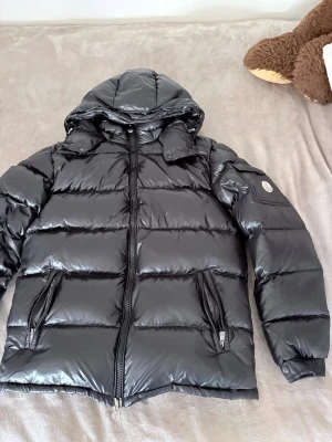 Svart dunjacka från Moncler - Snygg svart dunjacka från Moncler med glansig finish och huva. Jackan har två dragkedjeförsedda fickor framtill och en Moncler-logga på ärmen. Insidan har ett unikt serietidningsmönster och märkestaggar. Perfekt för kalla vinterdagar. Inget kvitto!