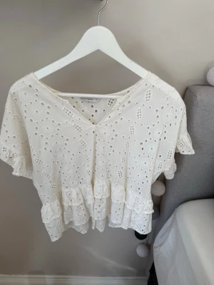 Vit broderad blus med volanger - Söt vit blus med broderade hålmönster och volanger nedtill. Blusen har v-ringning och korta ärmar med volangkant. Tillverkad i ett lätt bomullstyg som ger en luftig och romantisk känsla. Perfekt för en trendig och avslappnad look.