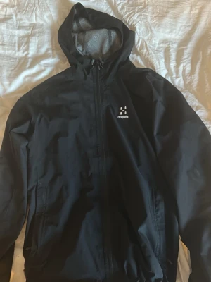 Svart vindjacka från Haglöfs Vårmodellen - Snygg svart vindjacka från Haglöfs med huva och hel dragkedja framtill. Jackan har meshfoder på insidan för extra ventilation och två sidofickor med dragkedja. Perfekt för blåsiga dagar och outdoor-aktiviteter.