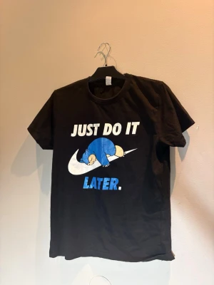 Svart nike t-shirt med Snorlax-tryck - Cool svart t-shirt med tryck av Snorlax som ligger på en Nike-liknande swoosh och texten 'Just do it later.' Trycket är i vitt och blått. Perfekt för dig som gillar Pokémon och vill ha en avslappnad stil.