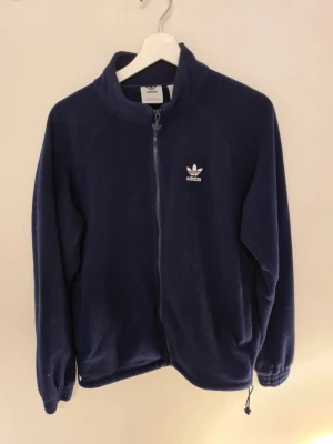 Mörkblå fleecejacka från Adidas - Mysig mörkblå fleecejacka från Adidas med dragkedja framtill och broderad logga på bröstet samt stor logga på ryggen. Jackan har hög krage och dragsko nertill för extra komfort. Perfekt för kyliga dagar.