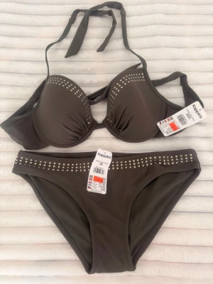 Mörkbrun bikini med nitar från KappAhl - Snygg mörkbrun bikini med vadderad bygel-bh och knyt i nacken. Både överdel och underdel har dekorativa silvriga nitar längs kanterna för en cool touch. Perfekt för strand eller poolhäng.