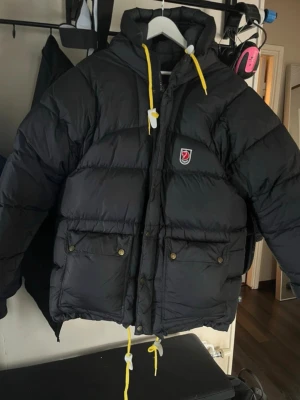 Svart fjällrävenjacka  - Säljer en svart dunjacka från Fjällräven med gula dragskor och luva. Jackan har två stora fickor framtill med knappar och ett klassiskt Fjällräven-märke på bröstet. Perfekt för kalla vinterdagar och riktigt varm tack vare dunfyllningen. Den har ett litet hål i nedre delen av jackan, som det finns bild på. Ett snöre är lite slitet men ingenting man tänker på, det finns också bild på.