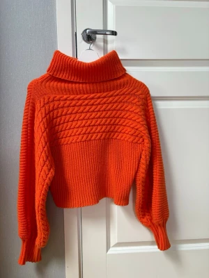 Orange polotröja med kabelstickning - Snygg orange stickad polotröja med breda ärmar och kabelstickat mönster över bröstet. Tröjan har en hög, vikt krage och ribbstickade muddar. Perfekt för att hålla värmen och sticka ut i mängden.