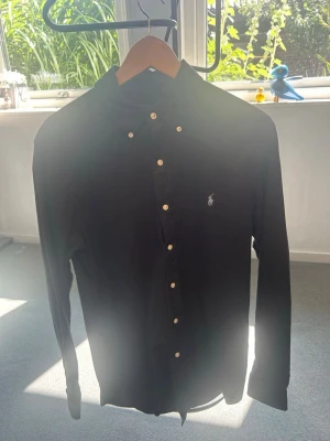 Svart skjorta med knappar från Polo Ralph Lauren - Klassisk svart skjorta från Polo Ralph Lauren med långa ärmar och vita knappar. Skjortan har button-down krage och den ikoniska loggan broderad på bröstet. Perfekt till jeans eller chinos för en clean och stilren look.