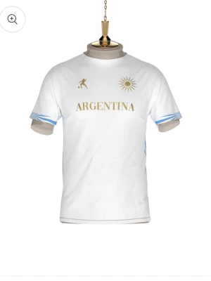 Vit Argentina fotbollströja - Snygg vit fotbollströja med guldiga detaljer och texten ARGENTINA på bröstet. Tröjan har korta ärmar med ljusblå mönster vid ärmsluten och tryck av en sol och en fotbollsspelare framtill. Perfekt för dig som älskar fotboll och vill sticka ut på planen. Och gör även byten med tröjor ifrån LAIB.