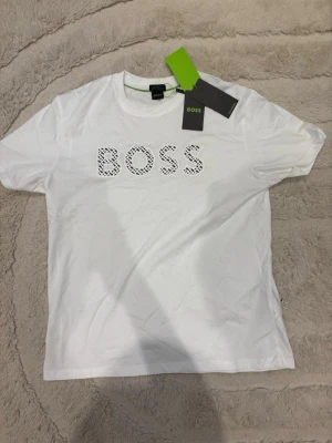 Vit t-shirt från BOSS  - Snygg vit t-shirt från BOSS med stor logga i svart och vitt mönster på bröstet. Klassisk rund halsringning och korta ärmar. Tillverkad i mjuk bomull för en bekväm känsla. Perfekt till jeans eller shorts för en clean look.