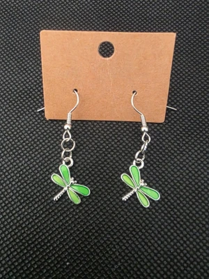 Gröna trollslända örhängen - Säljer ett par snygga örhängen med hängande trollsländor i grönt och silver. Örhängena har krokfäste och detaljerade vingar i emalj. Perfekt accessoar för dig som gillar naturinspirerade smycken och vill sticka ut lite extra.