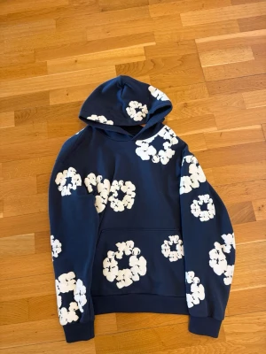 Denim tears hoodie - Snygg blå hoodie från Denim Tears med vita blomliknande tryck över hela tröjan. Tillverkad i 100% bomull och har en klassisk huva samt magficka. Perfekt för dig som vill sticka ut med en unik och trendig stil. Köpt på denim tears officiealla sida, Digitalt kvitto finns.