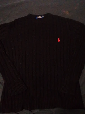 Svart kabelstickad tröja Polo Ralph Lauren - Kabelstickad svart tröja från Polo Ralph Lauren med klassisk röd logga broderad på bröstet. Tröjan har rund halsringning och ribbade muddar vid ärmslut och nederkant. Perfekt för dig som gillar stilrena och tidlösa plagg.