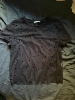 Svart topp med pärlor - Snygg svart topp med korta ärmar och rund halsringning. Toppen är dekorerad med små svarta pärlor över hela framsidan och baksidan för en extra cool look. Tillverkad i mjukt material som känns skönt mot huden.