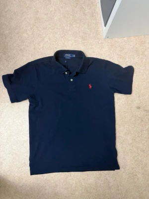 Marinblå pikétröja från Polo Ralph Lauren - Klassisk marinblå pikétröja från Polo Ralph Lauren med röd broderad logga på bröstet. Tröjan har korta ärmar, krage och knappar framtill. Perfekt för en clean och stilren look. I storlek M passar även bra för dom som har storlek S