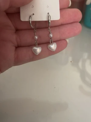 Silverörhängen med pärlhjärtan - Säljer ett par snygga silverfärgade örhängen med hängande vita pärlor och hjärtformade detaljer. Örhängena har en modern look med små pärlor ovanför hjärtat och passar perfekt för dig som gillar söta accessoarer.