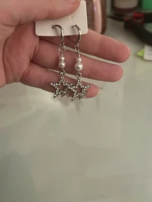 Silvriga stjärnörhängen med pärlor - Säljer ett par snygga hängande örhängen i silverfärg med stjärnformade hängen och vita pärldetaljer. Örhängena har en cool kedjedesign och passar perfekt för dig som gillar att sticka ut med accessoarer.