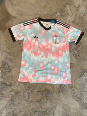 Pastellfärgad Adidas sporttröja S - Säljer en sportig t-shirt från Adidas i pastellfärger med rosa, blått och vitt mönster. Tröjan har svarta detaljer vid krage och ärmslut samt klassiska Adidas-ränder på axlarna. Tillverkad i ett lätt och ventilerande material, perfekt för träning eller sport.