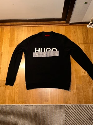 Svart sweatshirt från HUGO BOSS - Säljer en svart sweatshirt från HUGO BOSS med stor vit logga framtill och en grå detalj över bröstet. Tröjan har rund halsringning, långa ärmar och ribbade muddar vid ärmslut och nederkant. Perfekt för en stilren och avslappnad look.