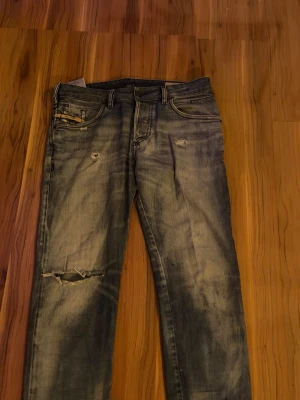 Grå slitna jeans med hål - Säljer ett par grå jeans med slitningar och hål på låret och vid fickorna. Jeansen har klassisk femficksmodell och raka ben. Perfekt för dig som gillar en avslappnad och trendig look.