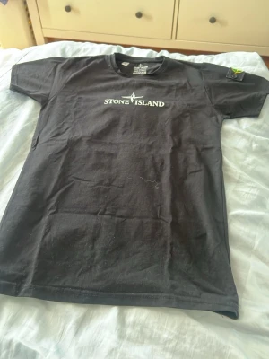 Svart Stone Island t-shirt - Säljer en svart t-shirt från Stone Island med klassisk logga på bröstet och den ikoniska patchen på ärmen. T-shirten har rund hals och korta ärmar, tillverkad i mjuk bomull för skön känsla. Perfekt för dig som gillar streetwear och vill ha något stilrent.