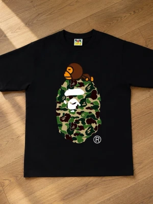  BAPE  - Svart t-shirt från BAPE med ikoniskt camo-mönstrat apansikte och en liten apa ovanpå. Klassisk passform och rund hals. Perfekt för dig som gillar streetwear och vill sticka ut med en unik design.