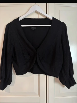 Svart kort kofta från Lindex - Kort svart kofta från Lindex med djup V-ringning och vida, långa ärmar. Koftan har en öppen front utan knappar och är gjord i ett mjukt, tunt material som ger ett elegant intryck. Perfekt att slänga över en topp för en snygg lager-på-lager-look. I storlek L.