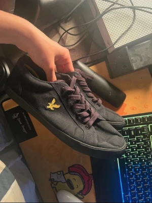 Lyle & scott skor  - Snygga svarta sneakers med tygöverdel och matchande svarta skosnören. På sidan finns en gul broderad logga i form av en fågel. Skorna har en klassisk låg profil och svart sula, perfekta för en clean och stilren look.
