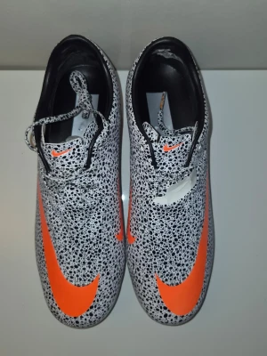 Nike fotbollsskor med orange swoosh - Säljer ett par Nike fotbollsskor med svartvitt prickigt mönster och stora orange swoosh-loggor. Skorna har snörning, låg profil och dobbar för spel på gräs. Insidan är svart och materialet är syntetiskt för bra grepp och kontroll på planen.