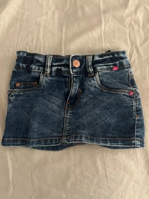 Mörkblå jeanskjol med fickor - Snygg mörkblå jeanskjol med klassiska fickor både fram och bak, bälteshällor och knapp framtill. Kjolen har kontrastsömmar och en liten rosa detalj vid fickan. Perfekt för en trendig och avslappnad look.