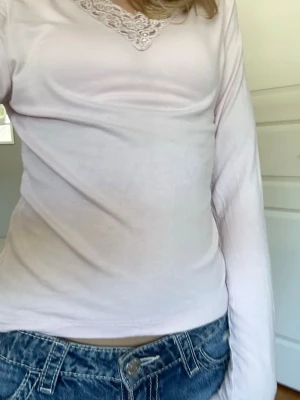 Ljusrosa långärmad topp med spets - Söt ljusrosa långärmad topp med v-ringning och spetsdetalj runt halsen. Toppen har en mjuk och bekväm passform, perfekt att styla med jeans eller kjol. Materialet känns lent och lätt mot huden.