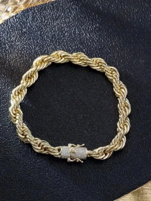 14k guldpläterad cordell armband - Stilrent armband i guldfärg med tvinnad design och ett lås dekorerat med små gnistrande stenar. Armbandet har en robust och elegant känsla, perfekt för att lyfta din look med en touch av lyx. Materialet ser ut att vara metall med glittrande detaljer på låset.