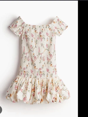 Blommig kortklänning med volang - Söt kortklänning i ljus färg med blommigt mönster och volang nedtill. Klänningen har offshoulder-korta ärmar och smockad överdel som ger en bekväm passform. Perfekt för varma dagar och somriga tillfällen.
