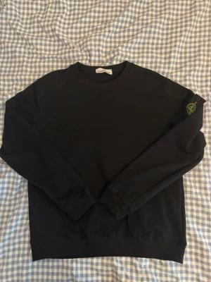 Svart sweatshirt från Stone Island - Säljer en svart sweatshirt från Stone Island med den klassiska loggan på vänster ärm. Tröjan har rund halsringning, långa ärmar och ribbade muddar vid ärmslut och nederkant. Perfekt för en clean och stilren look.