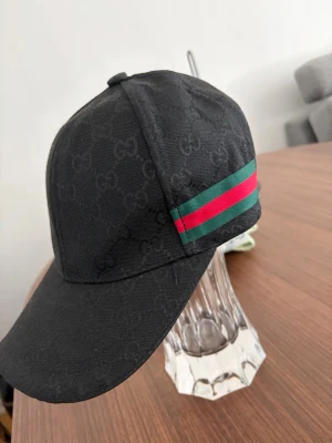 Svart  keps med-mönster - Snygg keps fråni i mörkbrunt tyg med klassiskt -monogram över hela kepsen. På sidan finns en grön och röd rand som ger den en ikonisk look. Perfekt accessoar för dig som vill sticka ut med stil.