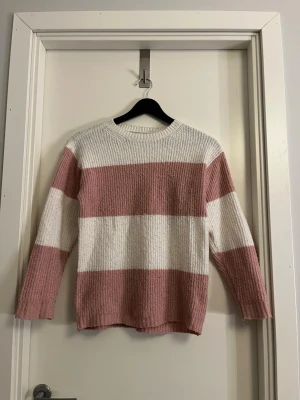 Randig stickad tröja från 157 - Mysig stickad tröja från 157 med breda ränder i vitt och ljusrosa. Tröjan har rund halsringning och långa ärmar. Perfekt för dig som gillar en avslappnad och trendig stil med mjukt material.